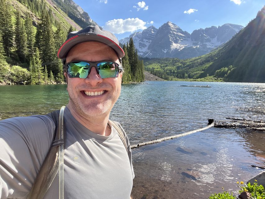 Jesse Thaler in sunglasses and a cap on a hike, with a mountain lake, pine forest and snow-capped peaks behind him